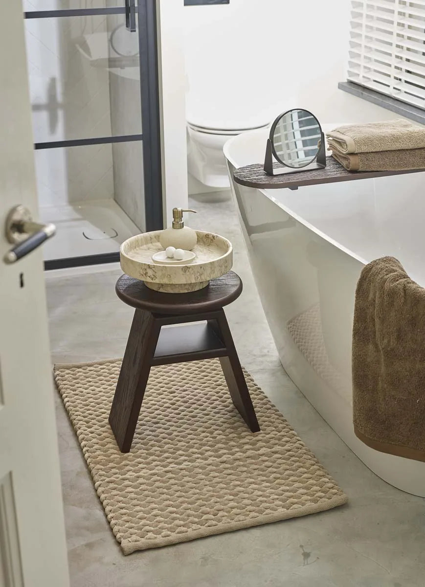 Badteppich in Beige mit strukturierter Oberfläche und dunklem Holzhocker neben einer weißen Badewanne
