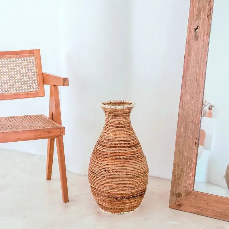Geflochtene Bodenvase Tumbak aus Bananenfaser neben Holzstuhl im Natur-Look.