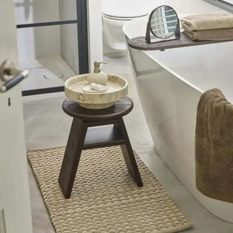 Badtrends 2027: Beiger Badteppich, dunkler Holzhocker und helle Badaccessoires neben einer weißen Badewanne
