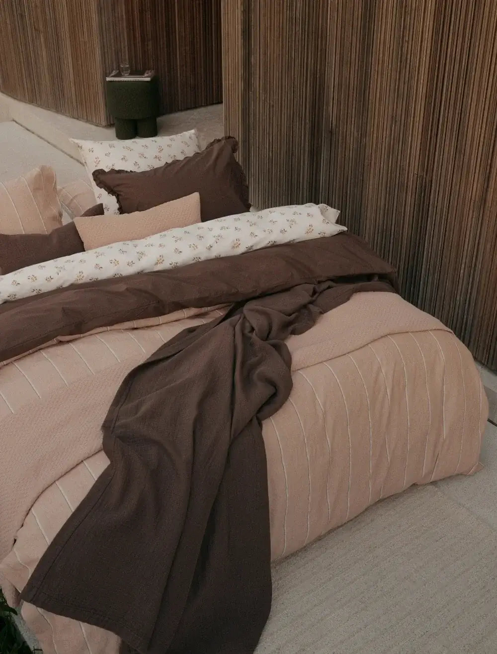 Tagesdecke Vandyck Pure 68 in Braun, locker über einem Bett mit rosabeiger Bettwäsche und gemusterten Kissen drapiert
