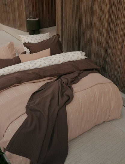 Tagesdecke Vandyck Pure 68 in Braun, locker über einem Bett mit rosabeiger Bettwäsche und gemusterten Kissen drapiert
