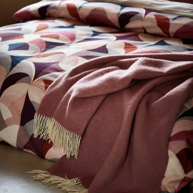 Wolldecke Vandyck Home Bellavita in Altrosa, Lammwoll-Plaid mit hellen Fransen auf einem Bett mit bunt gemusterter Bettwäsche
