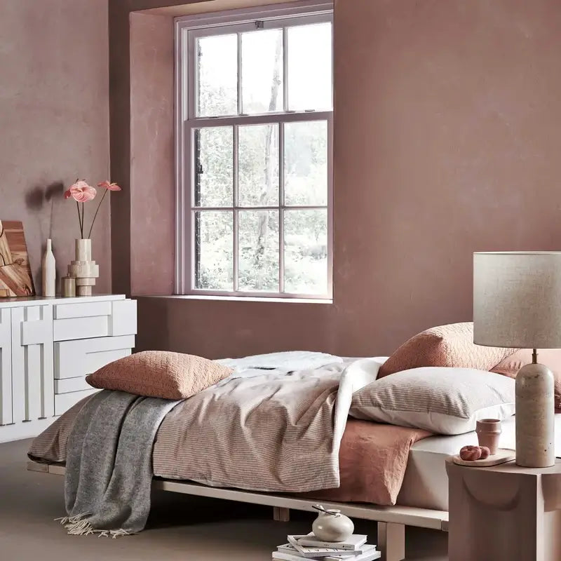 Wolldecke Vandyck Home Bellavita in Grau, Plaid aus Lammwolle mit hellen Fransen über einem Bett mit roséfarbener Bettwäsche
