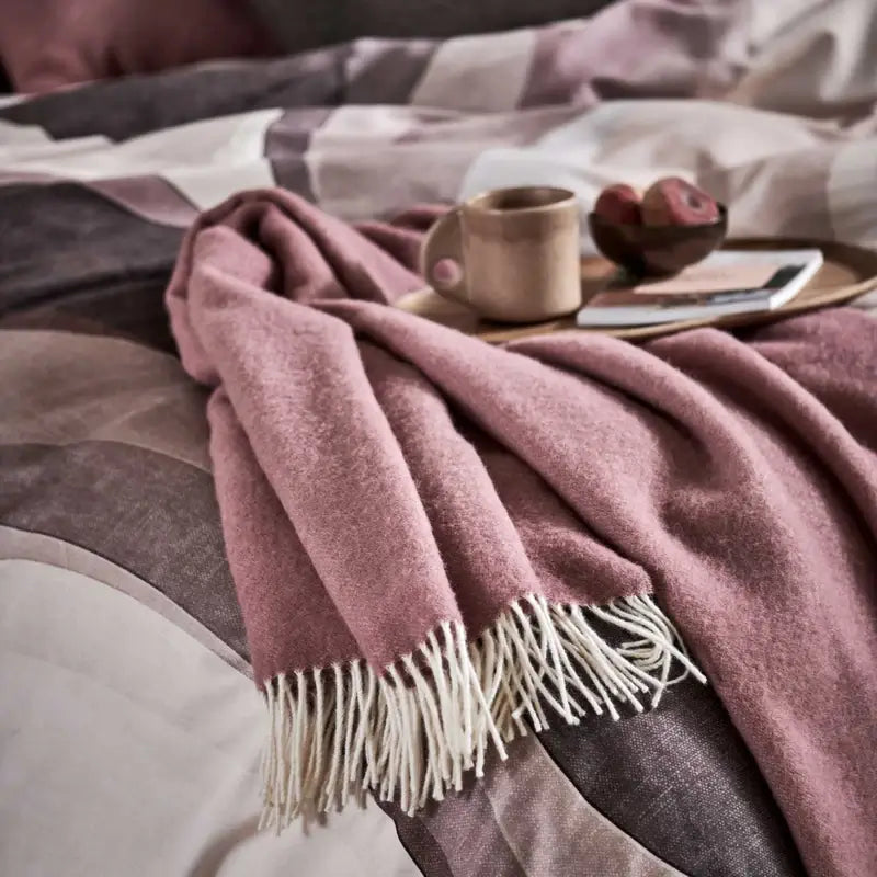 Wolldecke Vandyck Home Bellavita in Altrosa, Plaid mit hellen Fransen auf einem Bett neben einem Tablett mit Tasse und Buch
