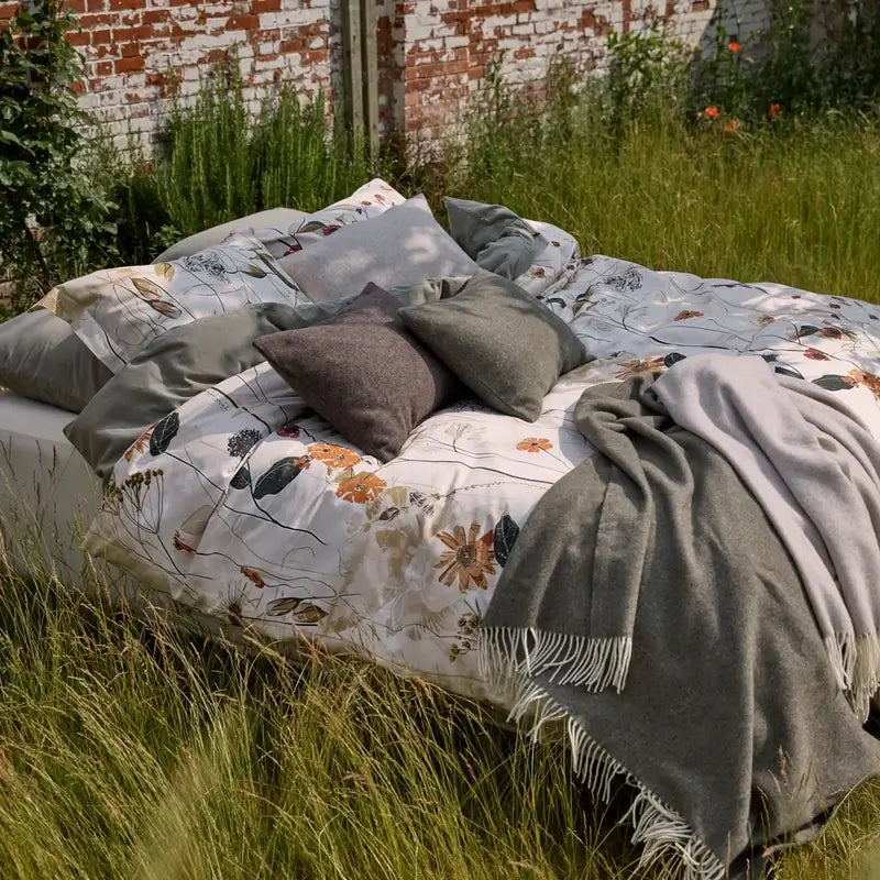Wolldecke Vandyck Home Bellavita in Grau, Plaid mit hellen Fransen auf einem Bett mit floraler Bettwäsche im Garten
