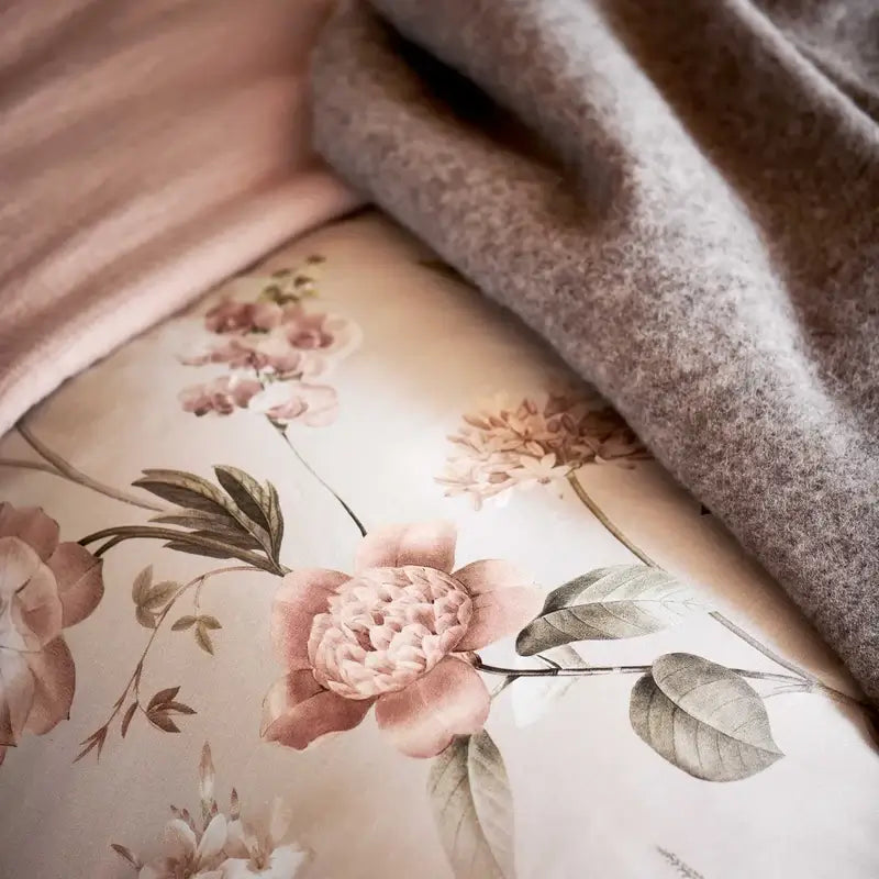 Wolldecke Vandyck Home Bellavita in Sand, Nahaufnahme des flauschigen Lammwoll-Plaids auf Bettwäsche mit rosa Blumen
